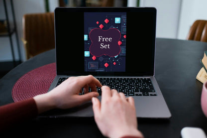 A person's hands typing on a laptop computer on a dark table. The laptop screen displays a digital graphic with the text 'Free Set' inside a purple frame, surrounded by abstract red and blue geometric shapes.